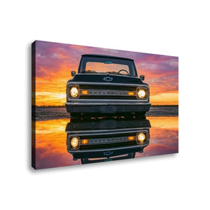 A square canvas wall art print showing a black 1970 Chevy C10 facing forward on a reflective surface at sunset, with a pink, orange, and purple sky. The canvas is photographed at a slight angle against a plain white background.