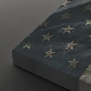 A close-up of a rectangular canvas or panel featuring a distressed American flag design, with a dark blue field and off-white stars, photographed on a dark gray background.