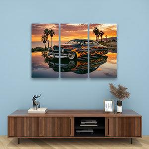 A three-panel canvas wall art print depicting a classic car with orange and yellow flames, parked in reflective water at sunset with palm trees, displayed above a wooden console table in a modern room setting.