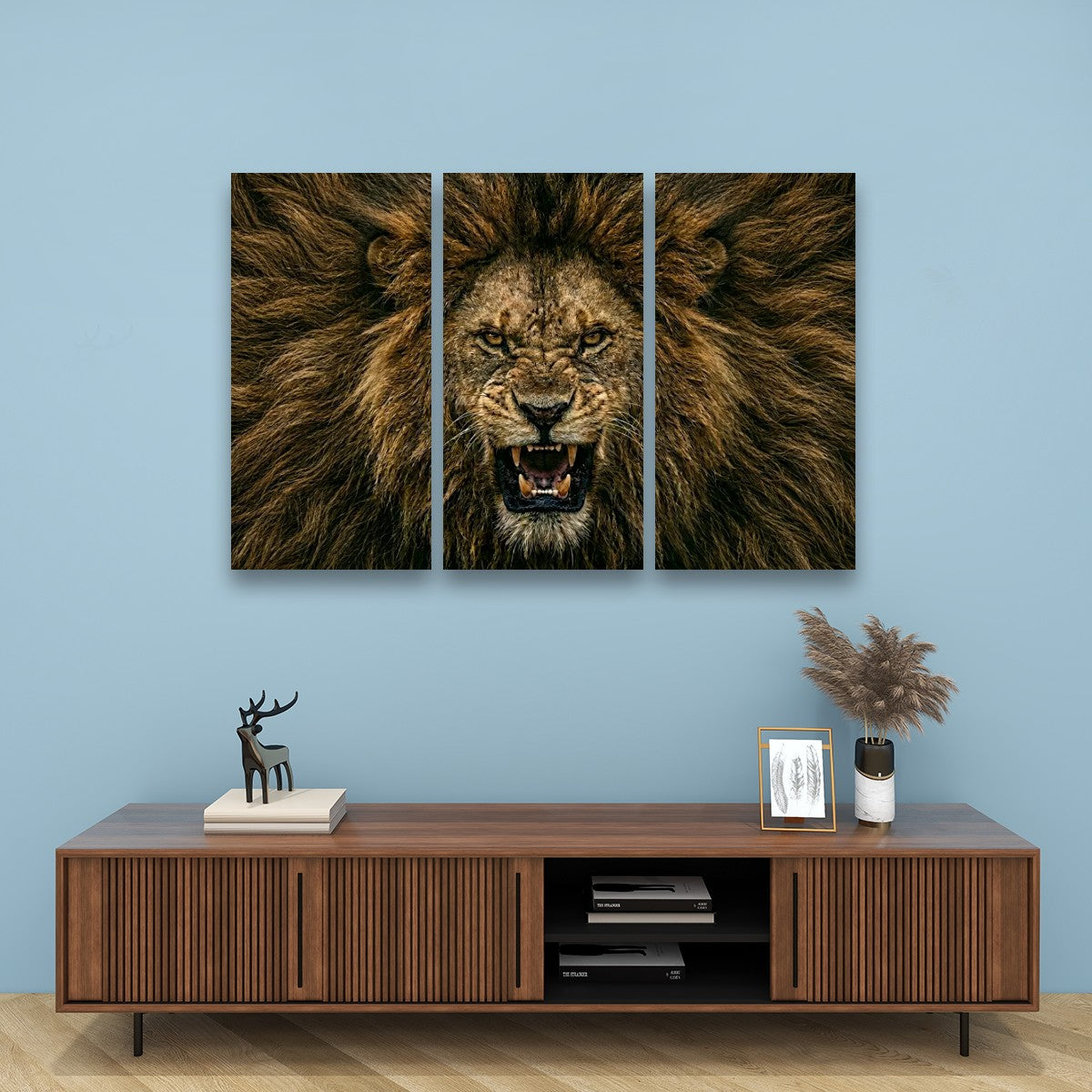 A three-panel canvas wall art set depicting a roaring lion's face and mane, displayed on a light blue wall above a wooden media console with decorative items.