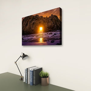 A rectangular wall art print showing a rocky coastline at sunset with bright orange and purple tones, displayed on a light wall above a desk with books, a lamp, and a small plant.