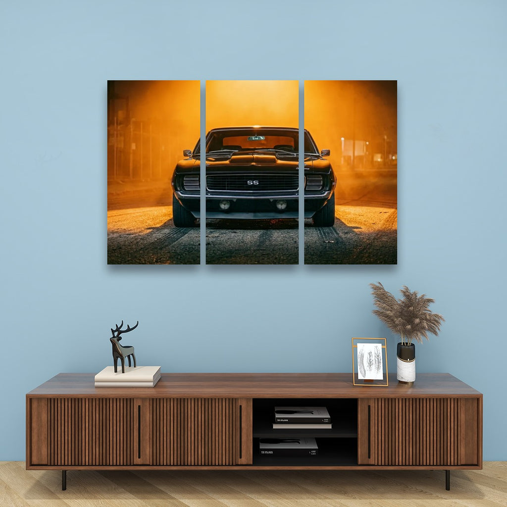 A three-panel canvas wall art print depicting a black 1969 RS/SS Chevy Camaro muscle car in a dusty, orange-lit environment, displayed on a light blue wall above a wooden console table with decorative items.