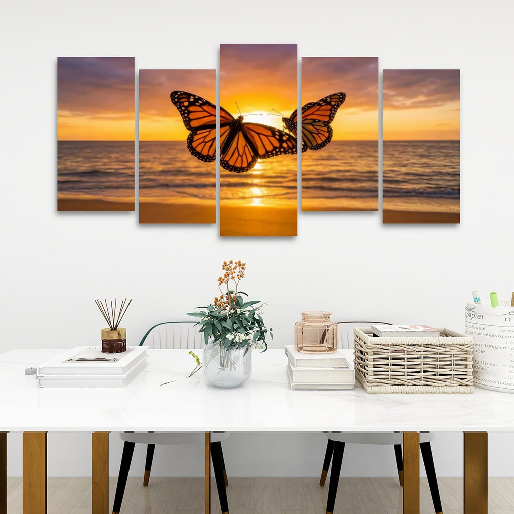 A 5-panel canvas wall art set depicting a monarch butterfly silhouette against a golden orange sunset over the ocean, displayed on a white wall above a styled dining table.