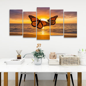 A 5-panel canvas wall art set depicting a monarch butterfly silhouette against a golden orange sunset over the ocean, displayed on a white wall above a styled dining table.