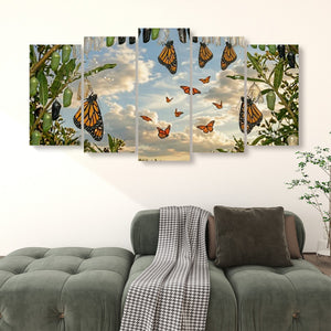 A five-panel canvas wall art set depicting monarch butterflies flying among green leaves and flowers under a cloudy sky, displayed above a green sofa in a living room setting.
