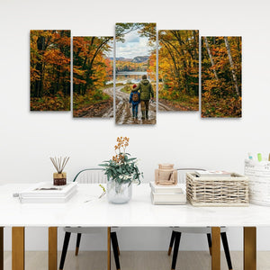 A five-panel canvas wall art set depicting a couple walking along a dirt road through an autumn forest with colorful fall foliage, displayed on a white wall above a styled dining table.