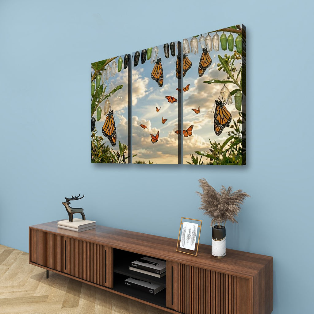 A three-panel canvas wall art print depicting monarch butterflies hanging from a tree against a cloudy sky, displayed on a light blue wall above a wooden console table with decorative items.