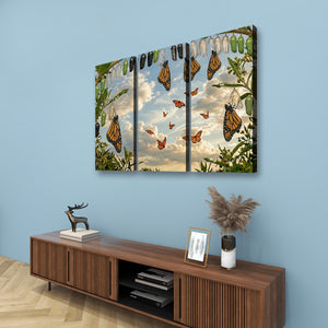 A three-panel canvas wall art print depicting monarch butterflies hanging from a tree against a cloudy sky, displayed on a light blue wall above a wooden console table with decorative items.
