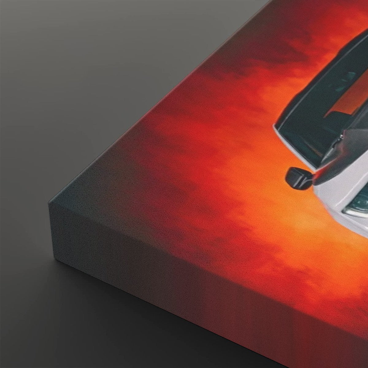 A close-up of a rectangular canvas print showing part of a white car against a fiery red and orange sky, photographed on a dark gray background.