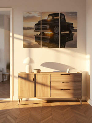 A three-panel canvas wall art print depicting a classic black 1956 Ford  F-100 pickup truck on a beach at sunset, displayed in a modern living room setting above a light wood sideboard.