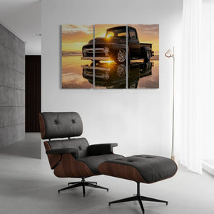 A three-panel canvas wall art print depicting a black vintage 1956 Ford  F-100 pickup truck on a reflective wet surface at sunset, displayed on a white wall in a modern living room setting above a brown and black lounge chair.