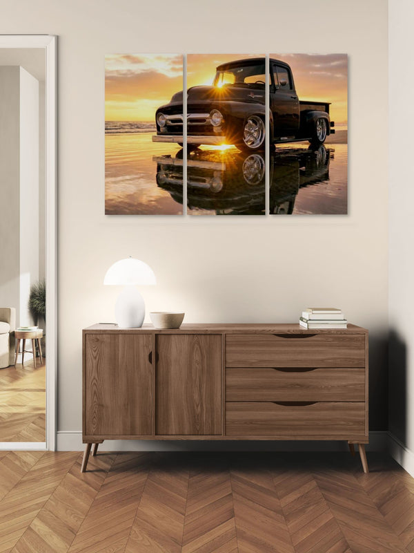 A three-panel canvas wall art print depicting a black vintage  1956 Ford  F-100 pickup truck at sunset with a reflection, displayed above a wooden sideboard in a modern room setting.