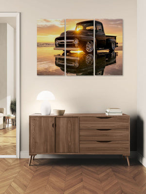 A three-panel canvas wall art print depicting a black vintage  1956 Ford  F-100 pickup truck at sunset with a reflection, displayed above a wooden sideboard in a modern room setting.