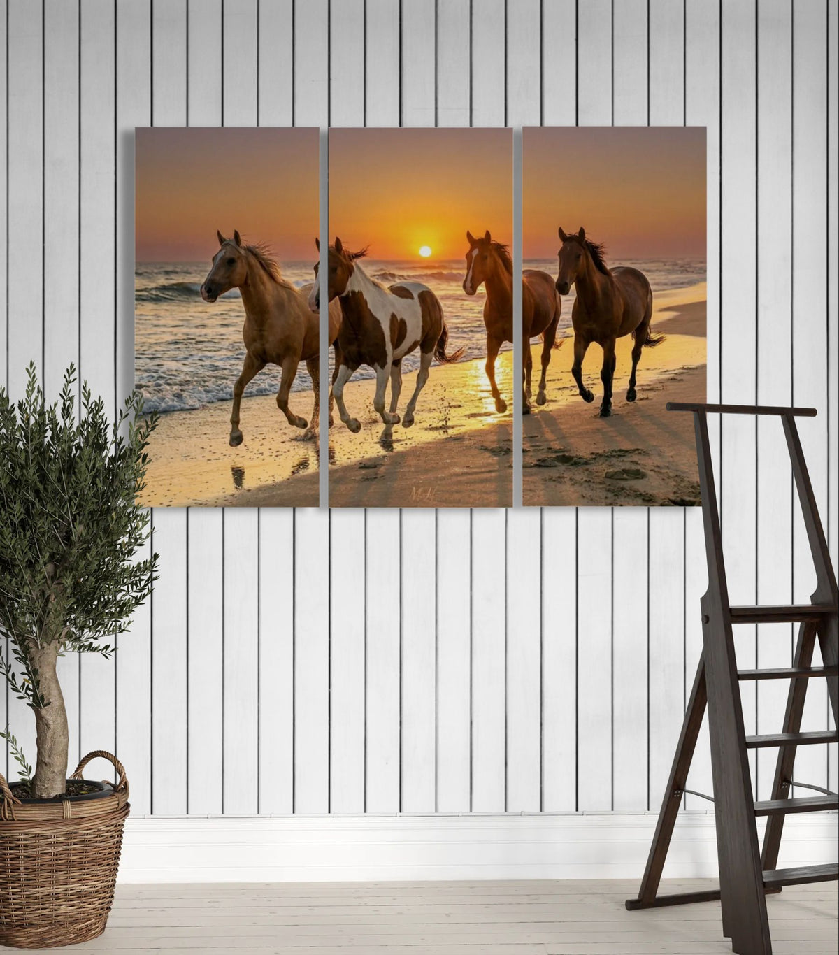 Triptych painting of horses on a beach at sunset hanging on a white wall.