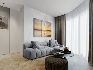 A modern living room featuring a gray sectional sofa, a round black coffee table, a dark woven ottoman, and a three-panel canvas wall art print depicting a beach sunset with a silhouette of a person walking on the sand.