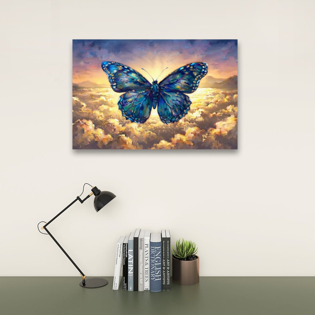 A rectangular wall art print showing a large blue butterfly over glowing clouds, displayed above a desk with a black lamp, books, and a small succulent against a light wall.