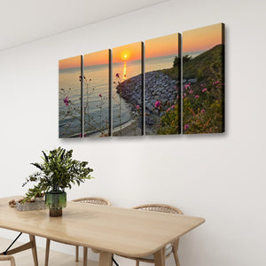 A four-panel canvas wall art set depicting a coastal sunset with rocks, flowers, and water, displayed on a white wall above a light wood dining table with chairs.