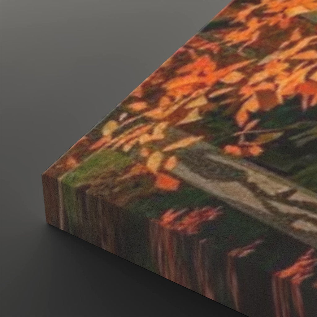 A close-up of a rectangular canvas or print showing an autumn landscape with orange and brown leaves, green grass, and a gray stone path, photographed on a dark gray background.