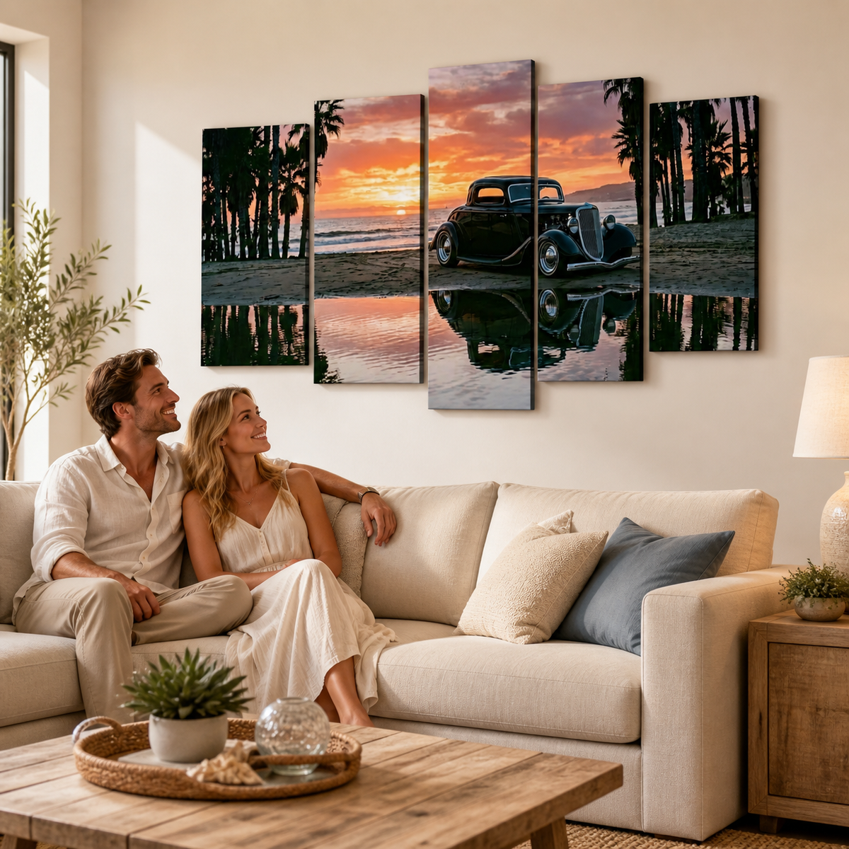 A couple sits on a cream sofa in a bright living room, looking at a five-panel canvas wall art print depicting a vintage 1934 Ford Coupe car on a beach at sunset with palm trees and water reflections.