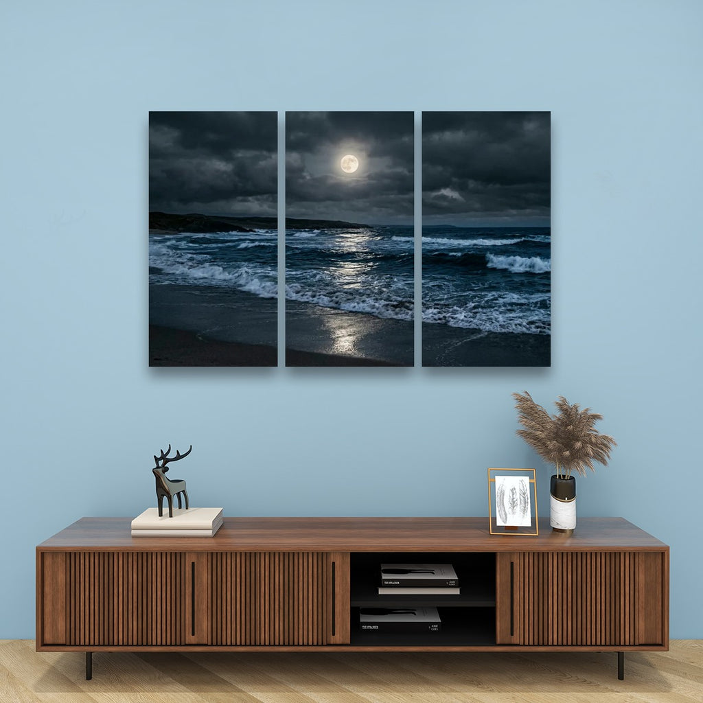 A three-panel canvas wall art set depicting a dramatic ocean scene at night under a full moon, displayed above a wooden console table against a light blue wall.