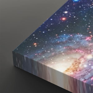 A close-up of a rectangular canvas or print showing a colorful galaxy or nebula scene, photographed on a dark gray background. The artwork contains many stars and glowing regions in blue, purple, pink, white, and warm orange tones.