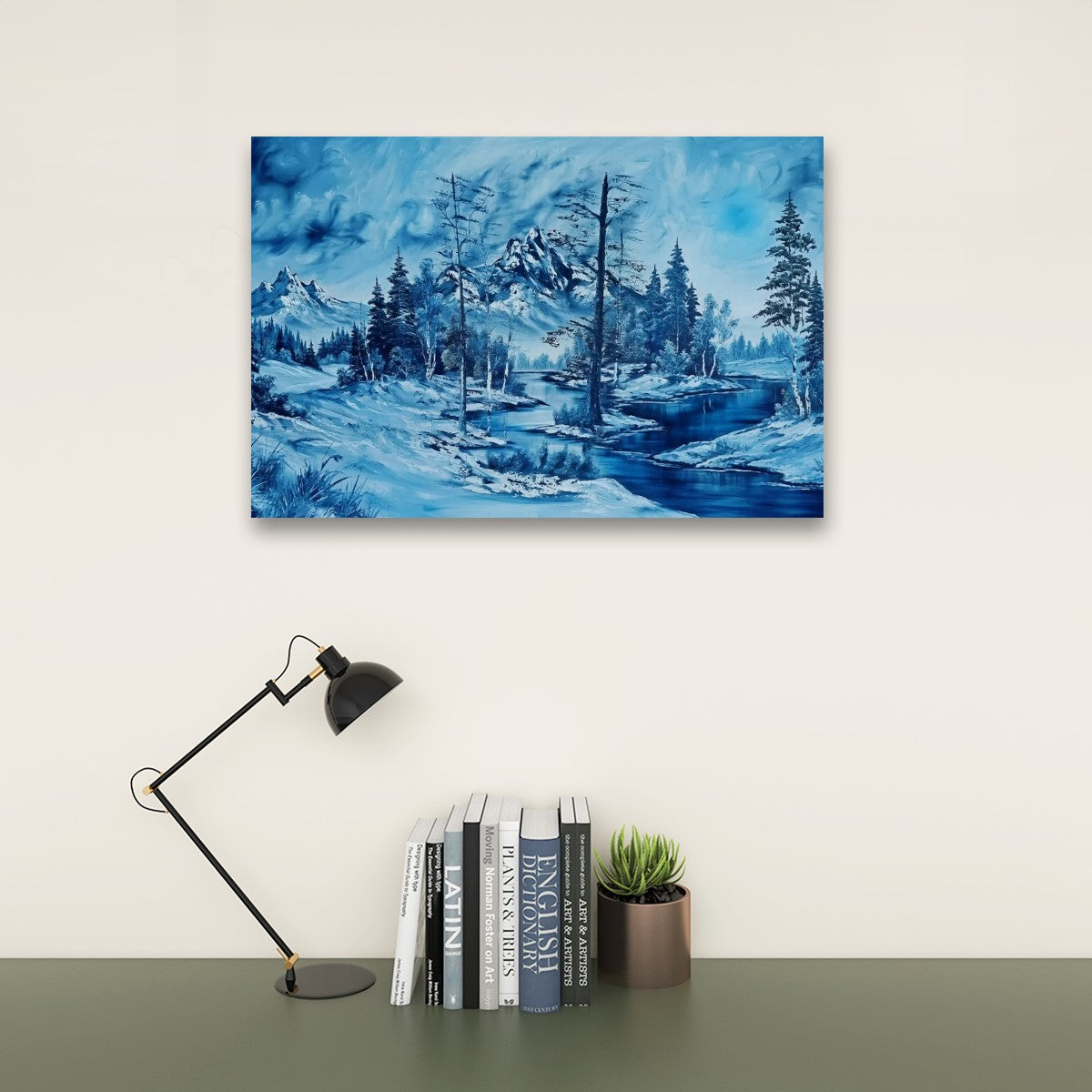 A rectangular wall art print showing a blue-toned winter landscape with snowy hills, trees, and a winding river, displayed above a desk with a black lamp, books, and a small potted succulent against a light beige wall.