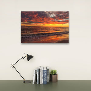 A rectangular wall art print showing a dramatic orange and red sunset over the ocean, displayed above a desk with a black lamp, books, and a small potted succulent against a light wall.