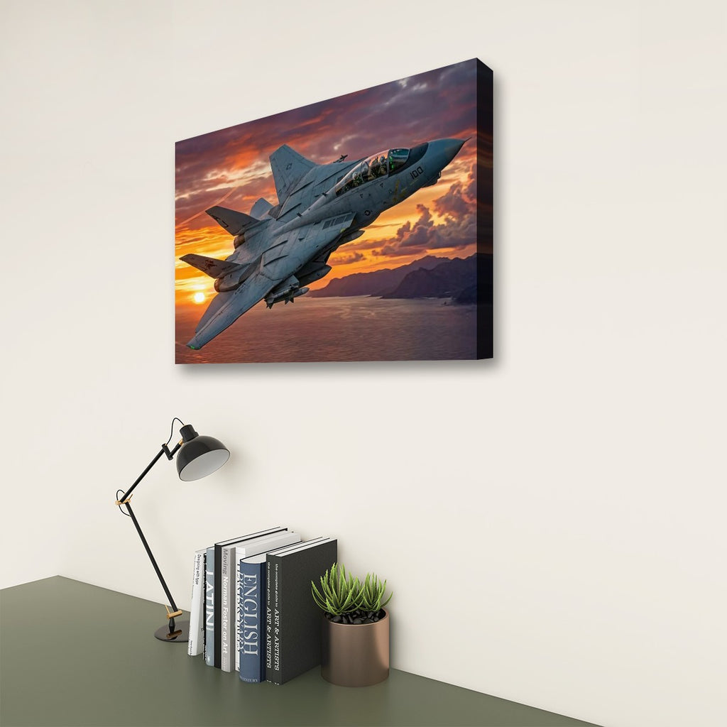 A rectangular wall art print showing a gray Grumman F-14 Tomcat  fighter jet flying over water at sunset, mounted on a light wall above a desk with a lamp, books, and a small plant.
