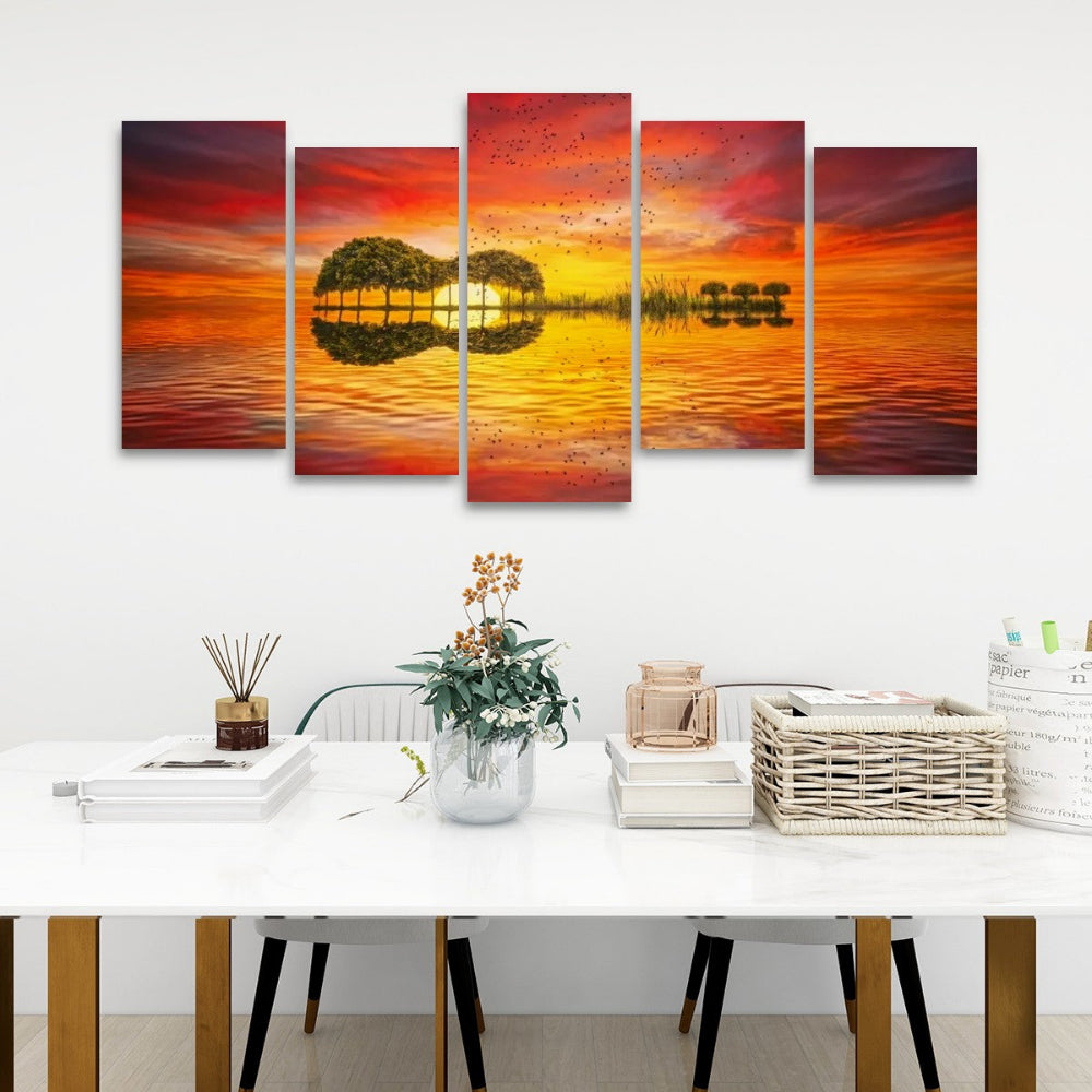 A 5-panel canvas wall art set depicting a sunset over water with tree silhouettes, displayed on a white wall above a styled dining table with books, a vase, and a basket.