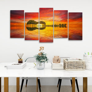 A 5-panel canvas wall art set depicting a sunset over water with tree silhouettes, displayed on a white wall above a styled dining table with books, a vase, and a basket.