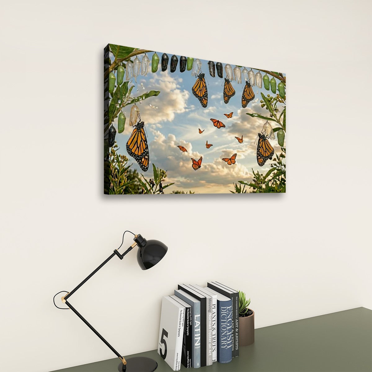 A rectangular wall art print showing monarch butterflies among green leaves against a blue sky with clouds, displayed above a desk with a black lamp and books in a styled interior setting.