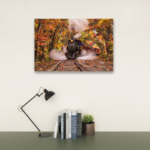 A rectangular wall art print showing a steam locomotive traveling through an autumn forest with colorful fall foliage, displayed above a desk with a black lamp, books, and a small succulent against a light wall.