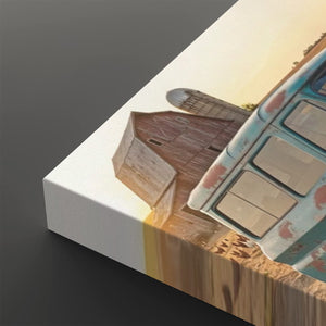 A close-up of a canvas wall art print showing a rustic barn and a vintage turquoise pickup truck in a desert landscape at sunset, photographed on a dark gray background.