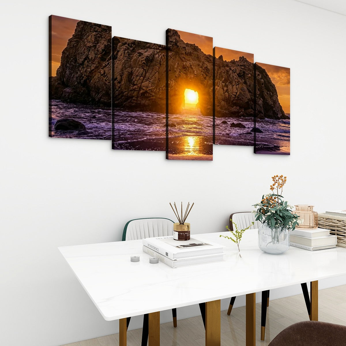 A five-panel canvas wall art set depicting a dramatic rocky coastline at sunset, displayed on a white wall above a modern white dining table with chairs and decorative items.