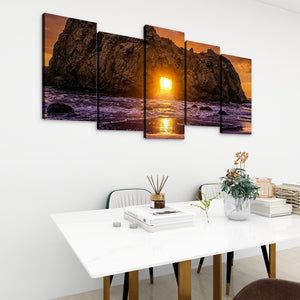 A five-panel canvas wall art set depicting a dramatic rocky coastline at sunset, displayed on a white wall above a modern white dining table with chairs and decorative items.