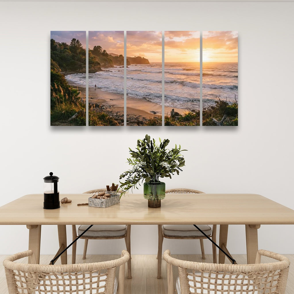 A five-panel canvas wall art set depicting a scenic sunset beach and ocean landscape, displayed on a white wall above a light wood dining table with chairs.