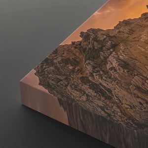 A rectangular canvas wall art print showing a rocky cliffside landscape under an orange sky, photographed at an angle on a dark gray background.