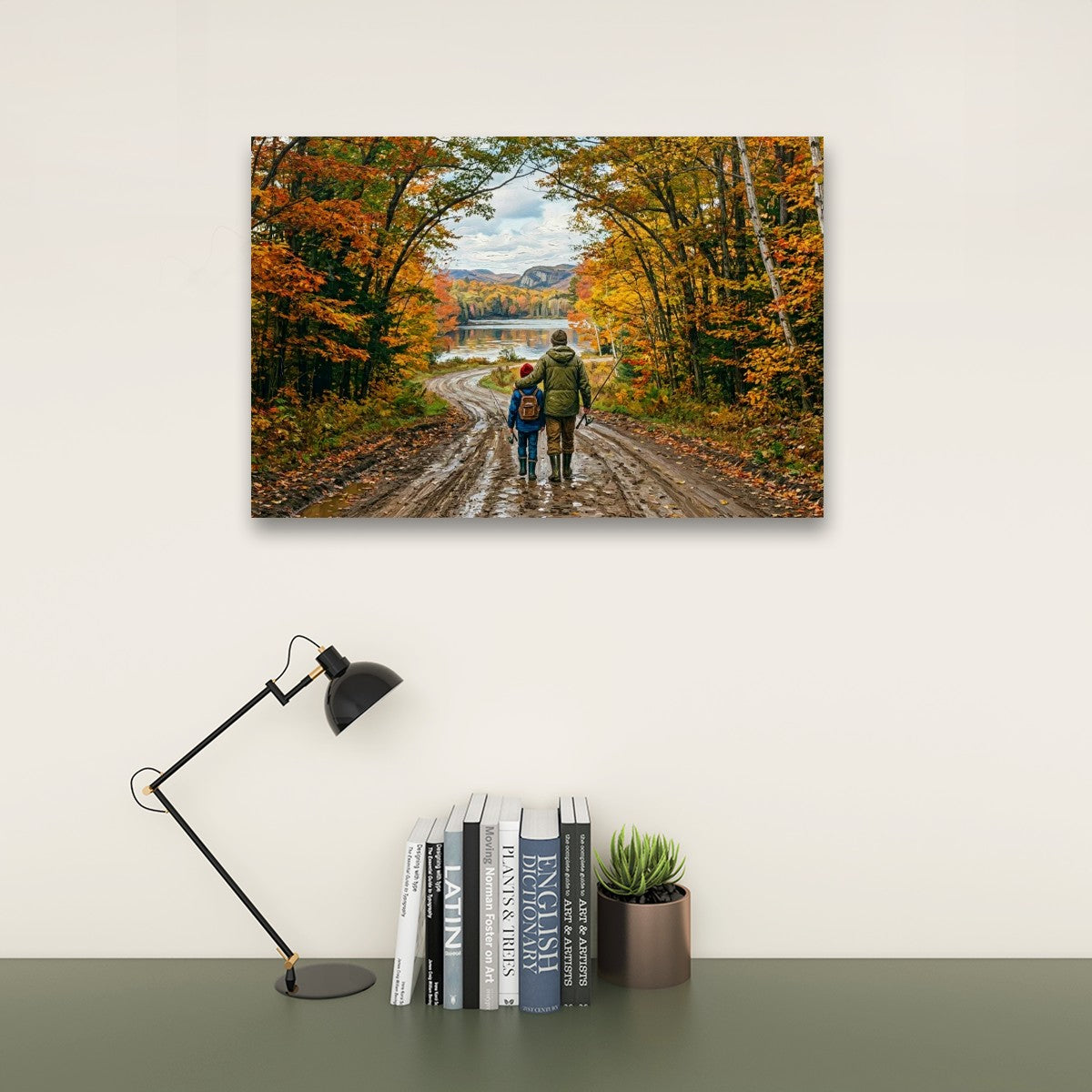 A rectangular wall art print showing two people walking along a dirt road surrounded by colorful autumn trees, displayed above a desk with a black lamp, books, and a small plant against a light wall.