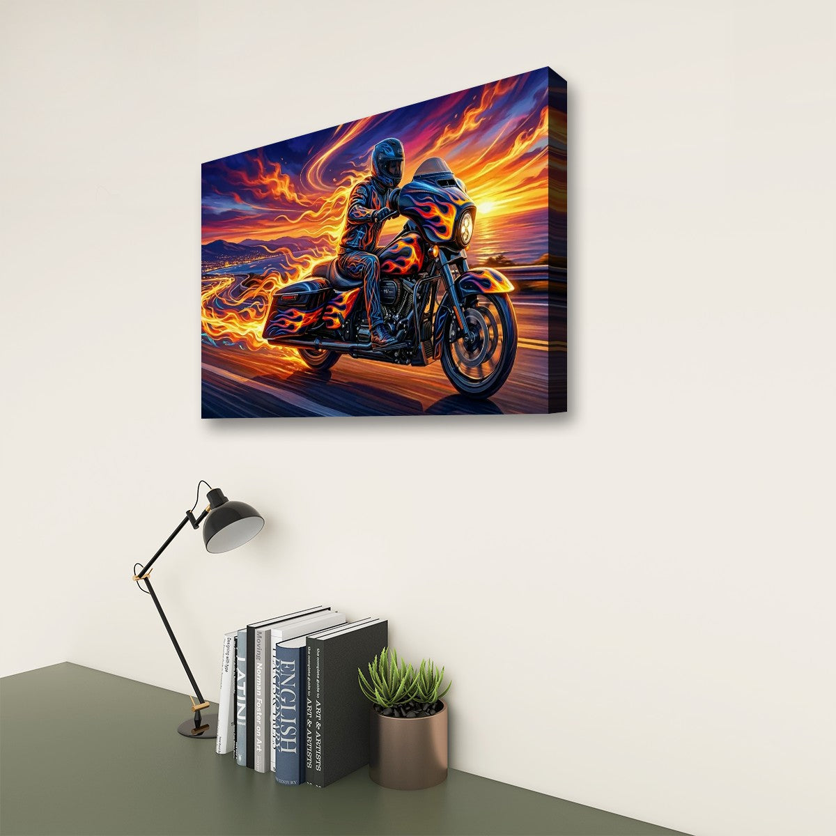 A rectangular wall art canvas showing a motorcycle rider on a road with dramatic orange, yellow, blue, and purple flames and sky, displayed above a desk with books, a lamp, and a small plant in a room setting.