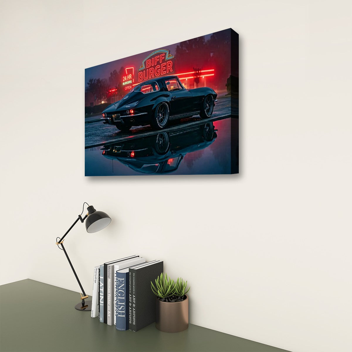 A rectangular wall art print showing a 1963 Split Window Chevy Corvette at night with red neon diner signage and a reflective wet road, displayed on a light wall above a desk with a lamp, books, and a small plant.