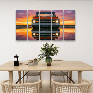 A multi-panel wall art print showing a vintage orange pickup truck facing forward against a vivid sunset reflection, displayed above a light wood dining table in a modern room setting.