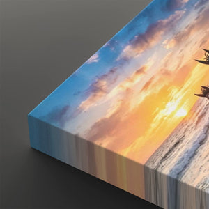 A rectangular canvas print showing a dramatic sunset over the ocean, photographed at an angle on a dark gray background. The artwork features a bright yellow and orange sun near the horizon, blue sky, and white sea foam, with the edge of the canvas visible on the left.