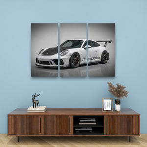 A three-panel canvas wall art print depicting a white Porsche 911 with black accents, displayed on a light blue wall above a wooden console table with decorative items.