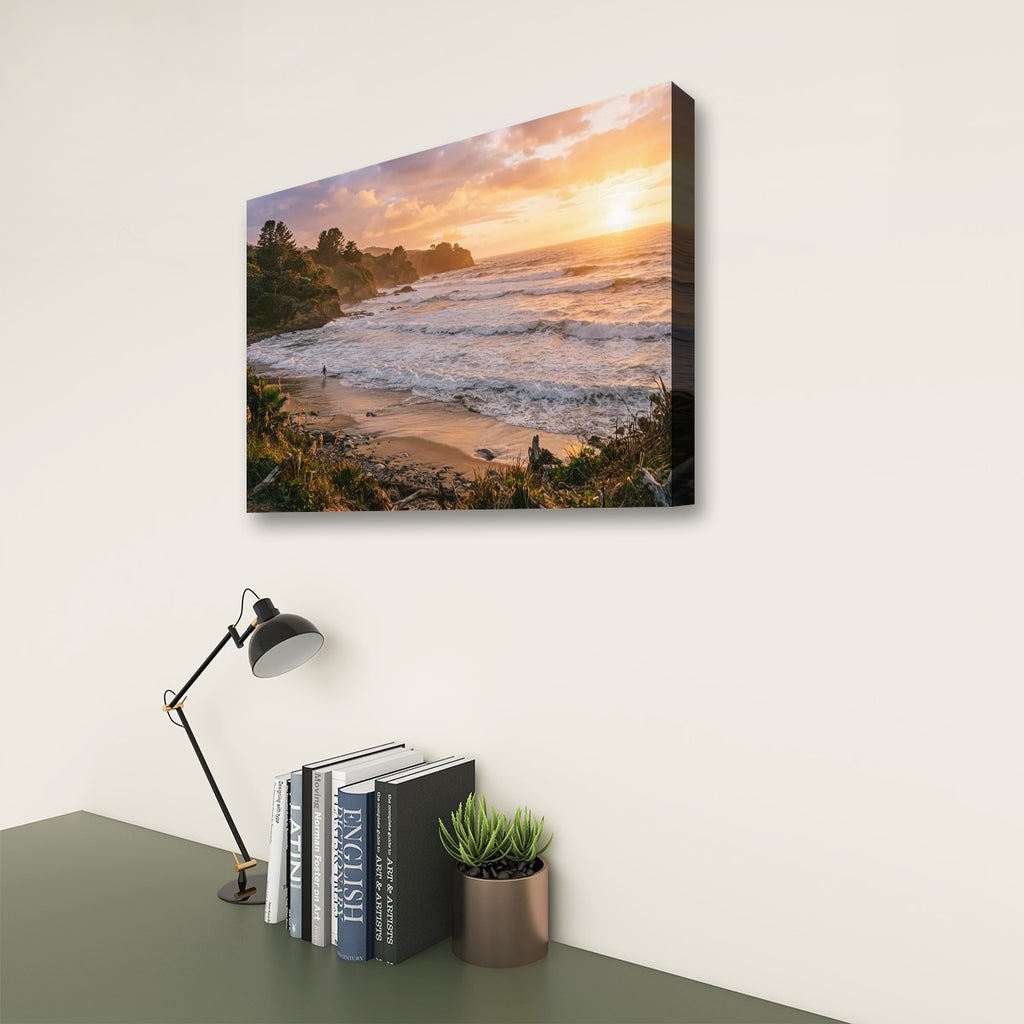A rectangular wall art print showing a beach and ocean at sunset, displayed above a desk with a black lamp, books, and a small potted succulent against a light wall.