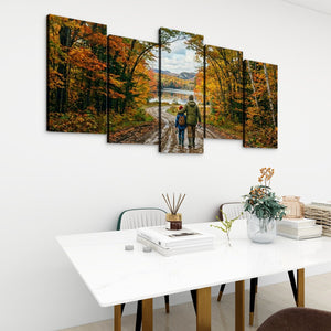 A 5-panel canvas wall art set depicting a couple hiking through an autumn forest with colorful orange and yellow foliage, displayed on a white wall above a modern white dining table.