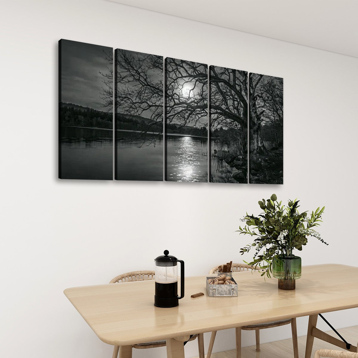 A five-panel black and white canvas wall art set depicting a moonlit lake scene with bare trees and moon reflection, displayed on a white wall above a light wood dining table with chairs and a plant.