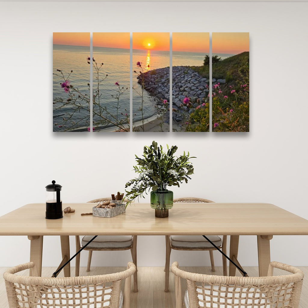 A five-panel canvas wall art print depicting a coastal sunset with flowers, displayed above a light wood dining table with woven chairs in a bright, modern room.