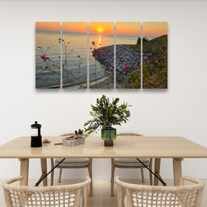 A five-panel canvas wall art print depicting a coastal sunset with flowers, displayed above a light wood dining table with woven chairs in a bright, modern room.