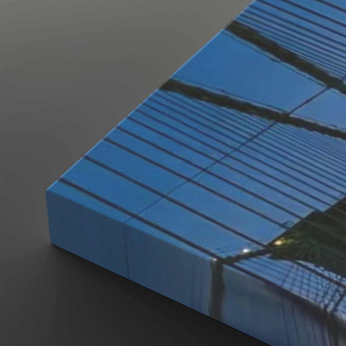 A rectangular canvas print or panel shown at an angle on a dark gray background, featuring a photographic print of a blue glass building facade with reflective lines and a small bright light reflection.
