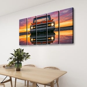 A four-panel canvas wall art print showing a red vintage pickup truck reflected in water at sunset, displayed above a light wood dining table in a modern room setting.
